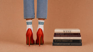 Weinsocken CIN CIN Winecraft, tealblaues Wording, rote High Heels, Weinbücher im Hintergrund.
