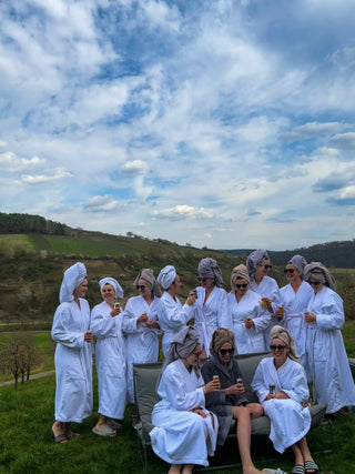12 Frauen in Bademänteln und Turbanen feiern Junggesellenabschied vor Bergpanorama mit Wein und Freundinnen